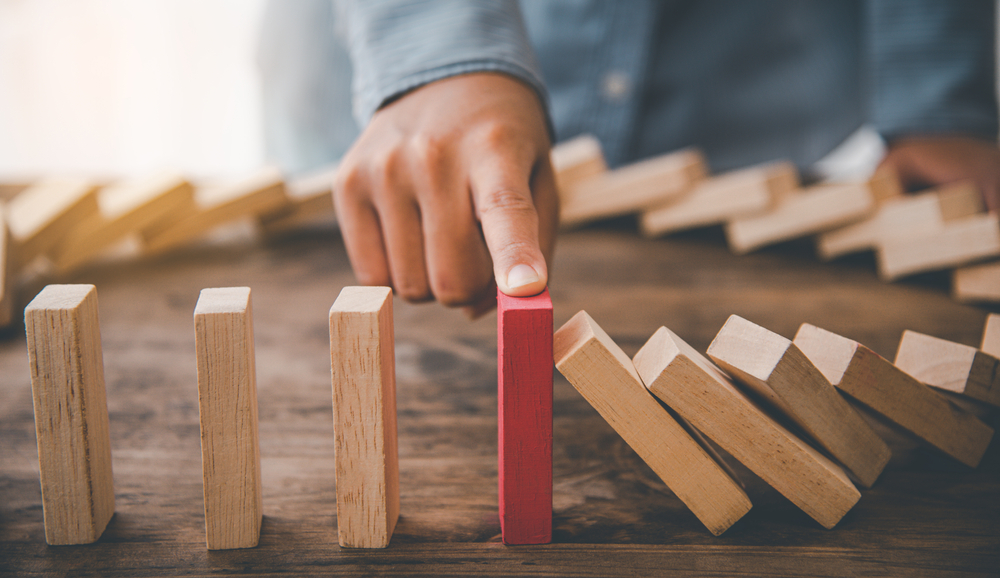 A hand stops a domino from tipping. Proactively preventing more dominions from tipping.